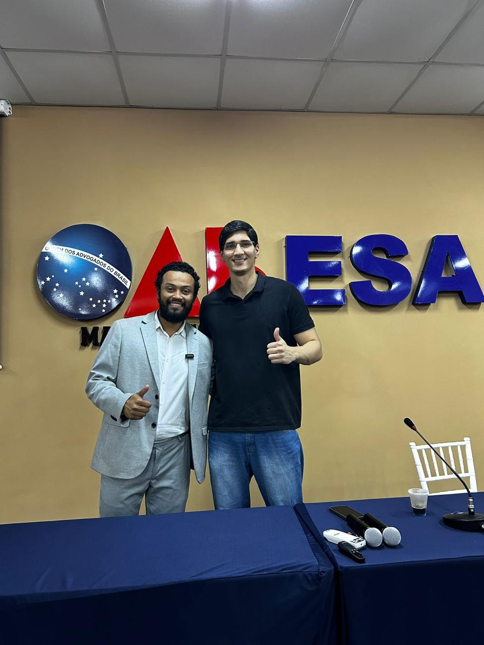 Dois participantes no espaço de eventos, em frente à identificação da OAB e da ESA.